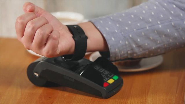 Close up shot of the payment process using smart watch. A person's hands produce a contactless plateade through a terminal in a small cafe, next to he a cup of coffee