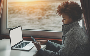 Serious curly female biracial teenager traveling by ship, sitting near window and sending message to her family while working on freelance job with modern laptop with white blank screen mock-up