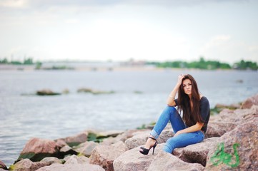Beautiful model in a black t-shirt, stylish jeans and shoes on high heels posing while sitting on stones near water