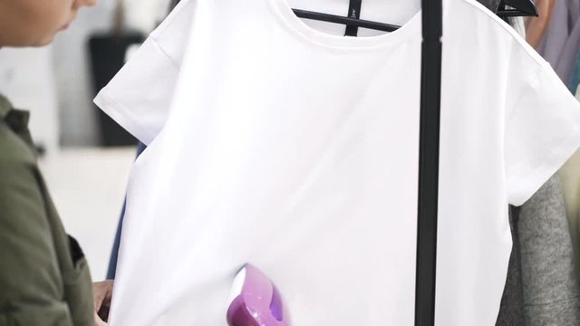Close up of a young woman wearing a green shirt steaming a white t shirt in a clothes shop. Handheld real time medium shot