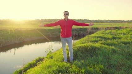 Young man in sunglasses with open arms is standing on the river bank at sunset