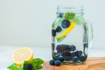 Infused detox water with lemon blackberry blueberry and mint leaves in a mason jar on a wooden board over white backgroun