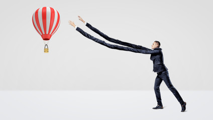 A businessman in side view trying to catch a large flying hot air balloon with his extra-long arms.
