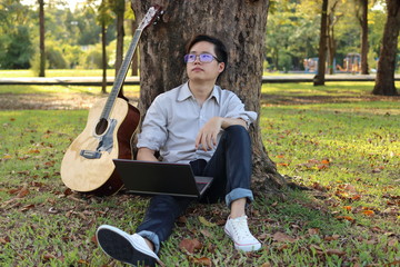 Happy hipster man leaning against a tree with a laptop and acoustic guitar. He looking far away in nature background.