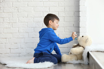 Cute little boy playing with toy bear and inhaler while sitting on floor near window. Allergy...