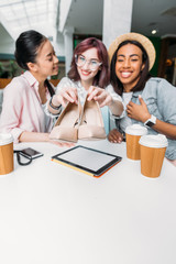 Smiling young women sitting at table with paper cups and showing new stylish shoes, young girls shopping concept