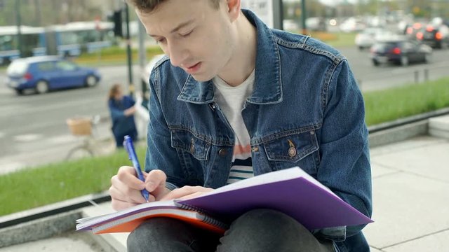 Young man looks absorbed while doing homework in the city, steadycam shot
