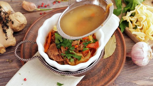 Pouring famous asian noodles soup - lamian - into white porcelain bowl on wooden kitchen table with soup ingredients - ginger, peppercorns, noodles, herbs. Top view.