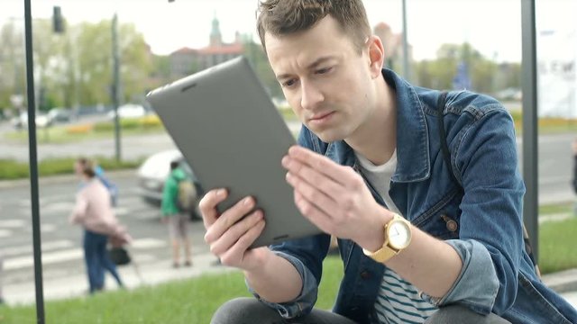 Young man squats in the city and smiling to the camera while using tablet, steadycam shot
