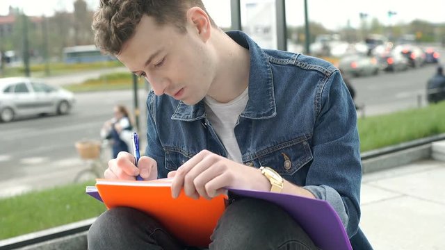 Young man smiling to the camera while doing homework in the city, steadycam shot
