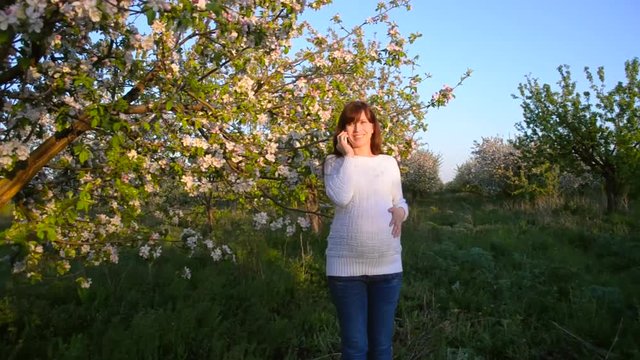 pregnant woman smiling talking on phone in flowering Park in spring, girl walks in blooming Apple orchard