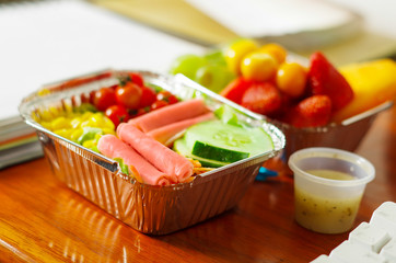 Delicious mediterranean-Style fresh salad with ham, with a blurred fruit salad on aluminium box on wooden table