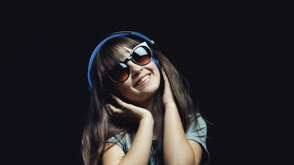 Studio shot of cute teenager girl wears headphones and sunglasses before black background, listening rhytmical music, youth freedom concept
