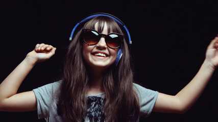 Studio shot of cute teenager girl wears headphones and sunglasses before black background, listening rhytmical music, youth freedom concept