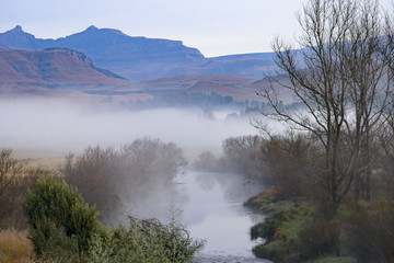 Fototapeta na wymiar A river runs through the foothills of the Drakensberg Mountain Range at Underberg in South Africa. This landscape-orientation takes in the mountains and a section of the Ngangwane River in the mist.