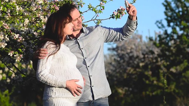 husband pictures of phone pregnant wife in blossomed spring Park, happy couple expecting child