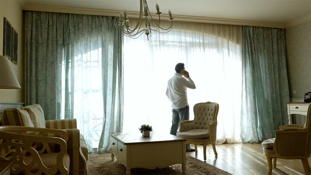 Worried man going forth and back along the window of a cozy apartment, talking on cell phone