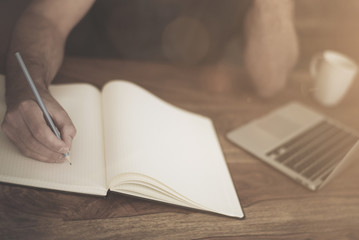 man at rustic wooden desk taking notes on pad while working on a laptop computer