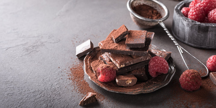 Broken chocolate pieces and raspberries with cocoa powder on metal plate on brown background with copy space. Dark photo.
