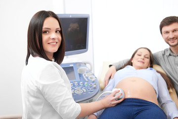 Obraz premium Husband embracing his pregnant wife by and carrying, when female doctor using ultrasound equipment diagnosing woman. Smiling doctor at modern hospital with patients making diagnostic procedure.
