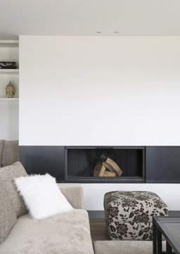 Salon moderne avec chemin&eacute;e encastr&eacute;e, canap&eacute; en tissu beige, coussin en fourrure blanche et pouf en tissu fleuri