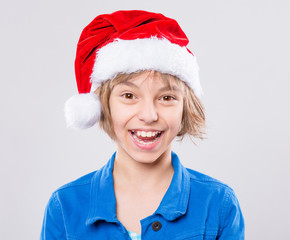 Emotional portrait of attractive caucasian little girl wearing Santa Claus red hat. Funny cute smiling child 10 year old looking at camera on gray background. Winter holiday christmas concept. 