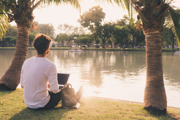 Young guy study while sitting on the grass by the water in a park. Warm tones in sunset. Copy space. Study or freelance working concept.