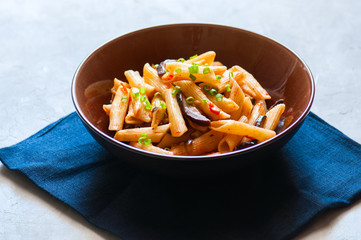 Penne pasta with egplants, tomatoes and green onions on a white stone background.