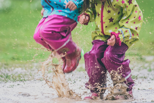 Children In Rubber Boots And Rain Clothes Jumping In Puddle.