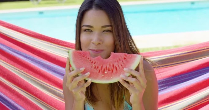 Cheerful young woman eating one watermelon slice while sitting on hammock stretched out behind her