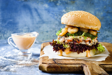 Asian style salmon burger with grilled shrimps, seaweed, lettuce and spicy sriracha mayo sauce served on pieces of brown paper on a rustic wooden board. Bright blue background with copyspace