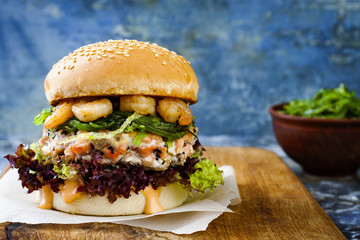 Asian style salmon burger with grilled shrimps, seaweed, lettuce and spicy sriracha mayo sauce served on pieces of brown paper on a rustic wooden board. Bright blue background with copyspace