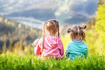 Fototapeta premium Two sisters sitting on the grass and look at the mountains. The concept of travel, lifestyle and family.