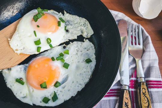 Fried eggs with green onion in pan, knife and fork on napkin, wooden background, top view