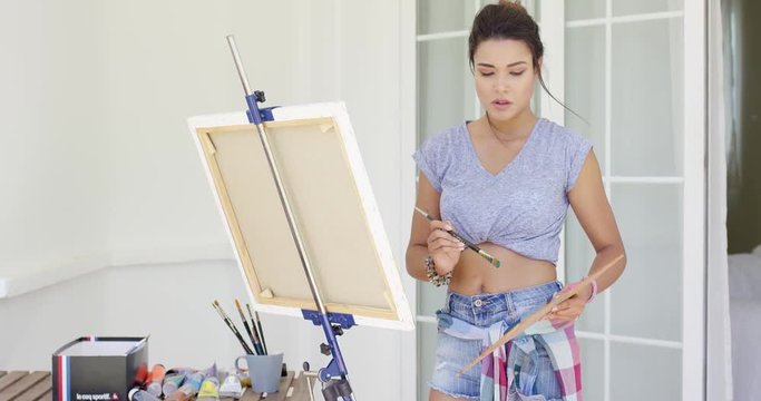 Attractive woman artist painting on her patio standing working on a canvas on an easel in summer shorts and bandanna