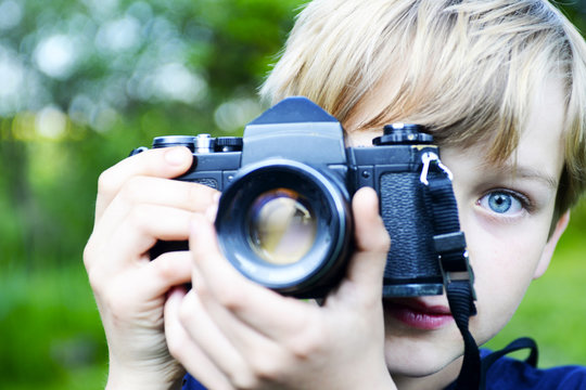 Little child blond boy with an old camera shooting outdoor. Kid taking a photo using a vintage retro film camera. 