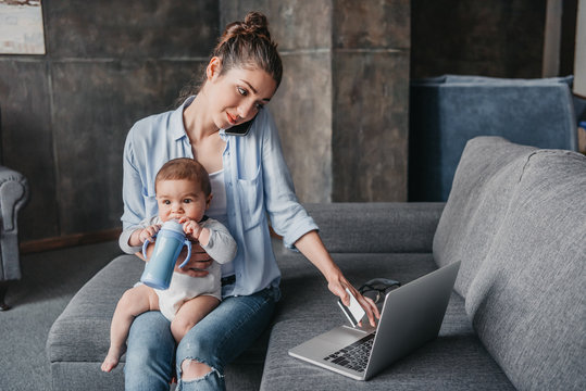 Young mother with baby boy remote working and using laptop during conversation on smartphone