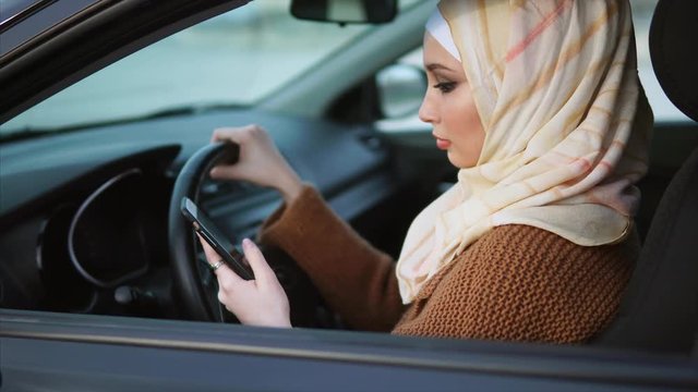 A modern pretty Muslim woman checking a notebook on her mobile phone during a traffic jam, a woman is driving a car. The simple life of an Arab woman in a big city