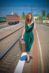 Gorgeous redhead woman in green velvet dress in vintage style with a suitcase on the platform