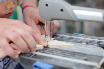 Dad Hands learning little son in protective glasses to fret saw board. Classes for young carpenter cutting wood form using fretsaw. Toy making. Dreaming of future profession. child woodcarving lessons