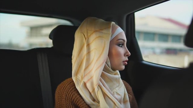 The modern life of a young Arab woman who is wearing a hijab, it rides in a car in the back seat and looks out the window. A Muslim with painted eyes goes to a meeting