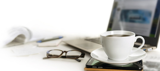 Coffee cup on an office desk with cell phone, laptop, glasses and papers, blurred background fades to white, panoramic banner for web page header, copy space - obrazy, fototapety, plakaty