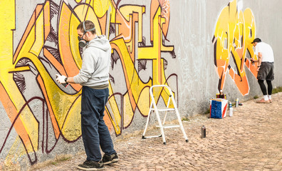 Urban street artists painting colorful graffiti on generic wall - Modern art concept with guys performing live murales with aerosol color spray - Focus on left person - Warm neutral filter