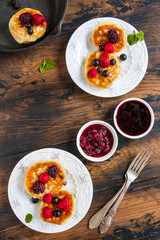 Homemade cottage cheese sweet pancakes with jam and berries, raspberry, black currant and blackberry. White ceramic plates, vintage forks, wooden rustic table, top view.