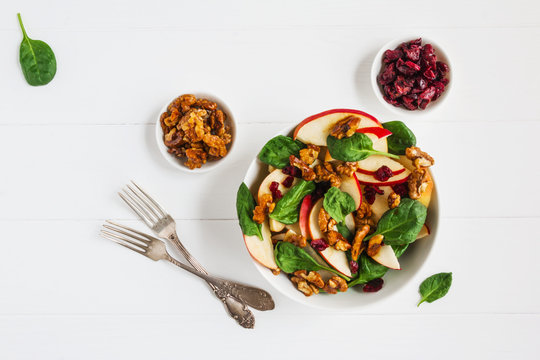 Healthy vegetarian salad with green spinach leaves, dried cranberry, red apple and walnuts caramelized in honey in a white bowl on the wooden table, top view.