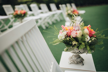 A composition of flowers and greens in a vase stands among the chairs for guests in the area of the wedding ceremony