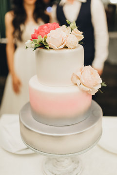 The two-level white wedding cake is decorated with flowers and stands on the banquet table on the background of the bride and groom
