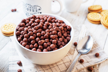 Hearty and tasty breakfast for children, cornflakes, chocolate balls with milk, cocoa and biscuits on a light wooden background 