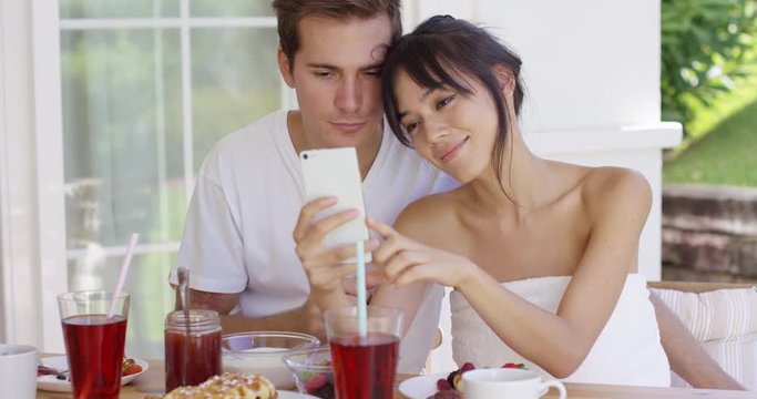 Smiling young woman taking picture of herself with attractive husband at table outside during breakfast time