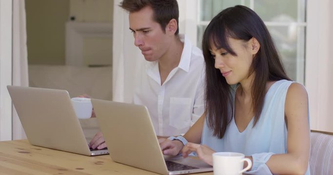 Calm young couple in loose summer clothing sitting at wooden table using similar laptop computers. Includes copy space.